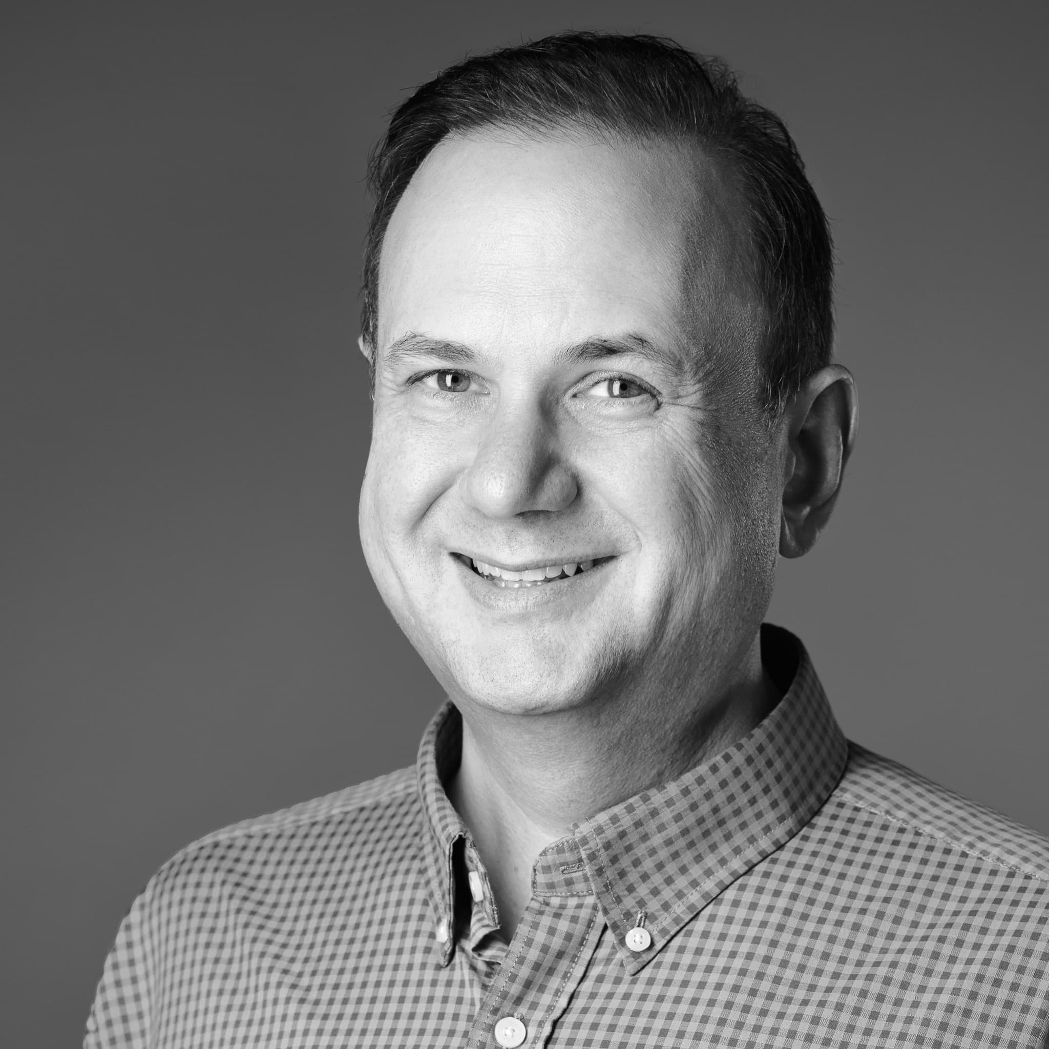 Black and white studio headshot in a gingham shirt against a gray backdrop.