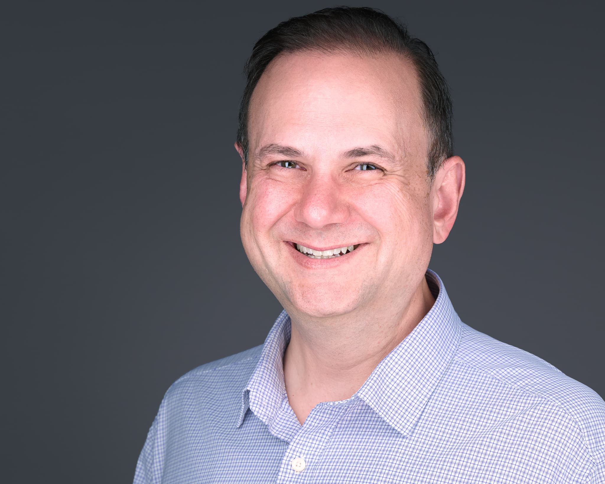 Color headshot in a light blue checked shirt against a slate backdrop, subject right of frame.