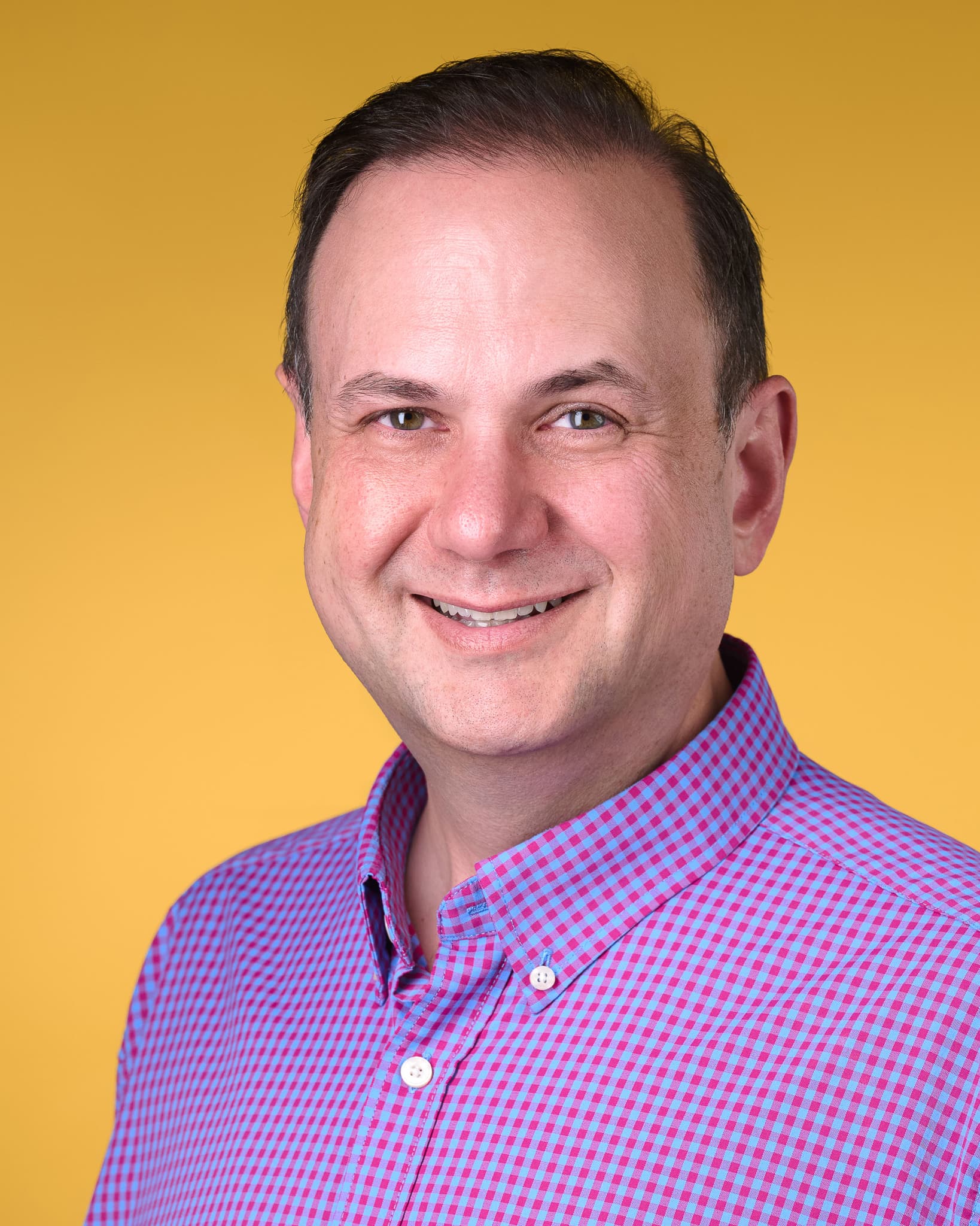 Color vertical headshot in a pink and blue gingham shirt against an amber backdrop.